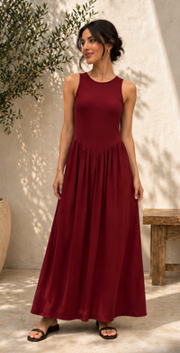 A woman wearing a sleeveless, floor-length maroon maxi dress with a gathered waist, standing in front of a textured beige wall with leaf shadows.