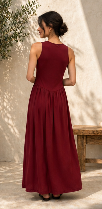 A woman wearing a sleeveless, floor-length burgundy maxi dress, shown from the back in an outdoor setting with a textured beige wall and soft shadows.