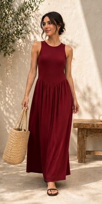 A woman wearing a sleeveless, high-neck, floor-length maxi dress in deep burgundy, holding a woven straw tote bag. She is standing outdoors against a textured beige wall with soft shadows, next to a rustic wooden bench.