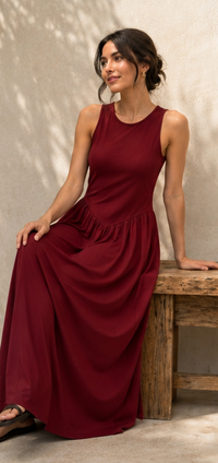 A woman wearing a sleeveless, floor-length burgundy maxi dress sits on a rustic wooden bench against a textured beige wall.