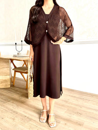A woman models a dark brown two-piece outfit consisting of a solid midi dress and a matching sheer open-front cardigan with wide sleeves, styled with white strappy heels and a woven straw bag in a bright indoor setting.