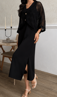 A woman wearing a black sheer mesh long-sleeve top with gold buttons over a black midi skirt with a front slit, paired with white strappy heels, in an indoor setting.