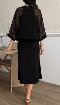 A woman shown from the back wearing a black sheer lace three-quarter sleeve top and a black midi skirt, standing in an indoor room with a light-colored wall and wood floor.