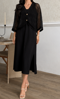 A woman models a black midi dress with a sheer black lace flutter jacket. The dress features a V-neckline, a front button closure with gold-toned buttons, and a side slit at the hem.