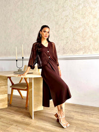A woman models a dark brown two-piece outfit consisting of a sheer patterned long-sleeve top and a matching wide-leg skirt, standing in a modern room with a light wood table and textured wallpaper.