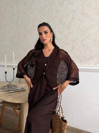 A woman models a dark brown two-piece outfit consisting of a sheer patterned short-sleeve cardigan and a matching dark brown satin slip dress, standing in a room with a textured wall and a wooden table.
