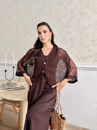 A woman models a dark brown two-piece outfit consisting of a matching brown slip dress and a sheer open-knit cardigan with textured patterned panels, holding a woven straw bag in an indoor setting.