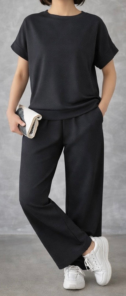 Person wearing a dark charcoal outfit with white sneakers against a gray background