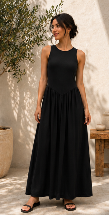 A woman wearing a sleeveless black maxi dress with a gathered waist and flowing skirt, standing in a sunlit outdoor setting with a textured wall, potted plant, and small wooden table.