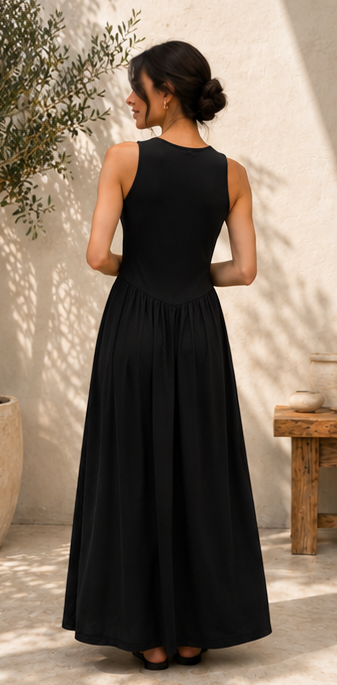 A woman wearing a sleeveless black maxi dress, shown from the back in an outdoor patio setting with a textured beige wall and soft shadows.