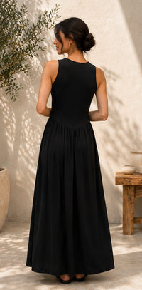 A woman wearing a sleeveless black maxi dress, shown from the back in an outdoor patio setting with a textured beige wall and soft shadows.