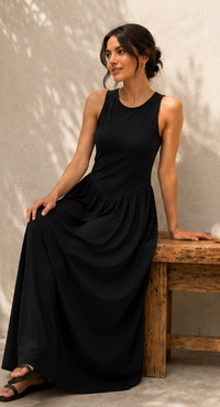 A woman wearing a sleeveless black maxi dress sits on a rustic wooden bench against a textured beige wall with soft leaf shadows.