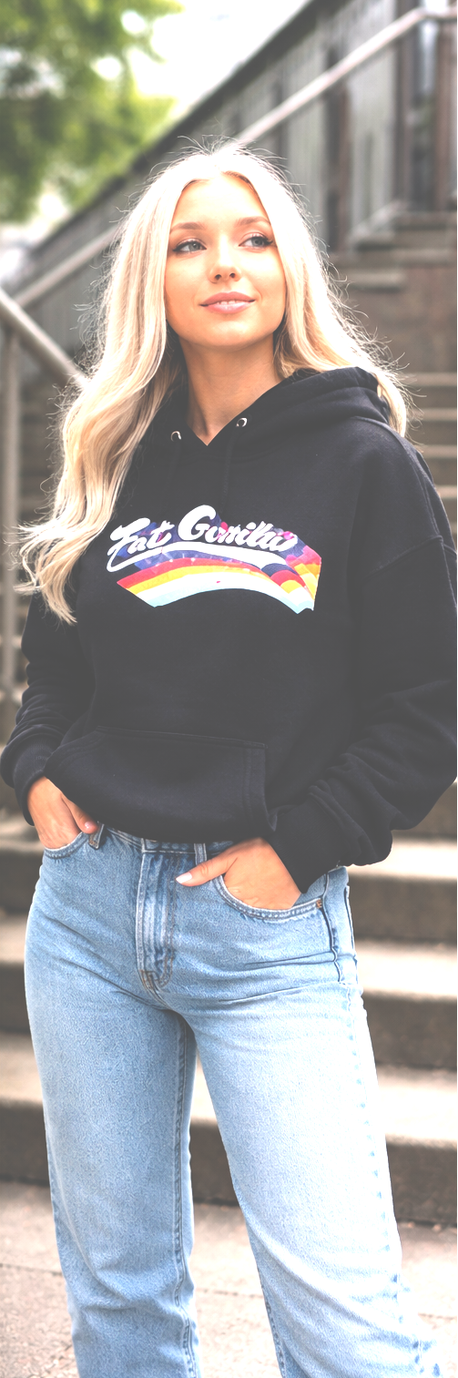 A woman wearing a black hoodie with a colorful 'Be Smitty' graphic on the chest, paired with light blue jeans, standing outdoors on steps.