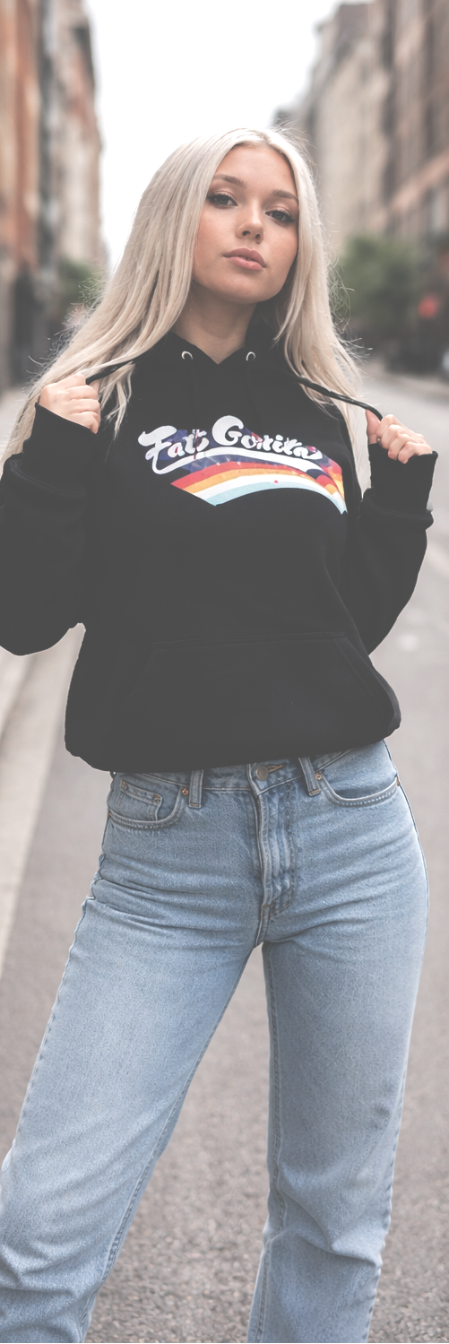 A blonde woman wearing a black hoodie with a white and red graphic on the chest, paired with light blue jeans, standing on a city street.
