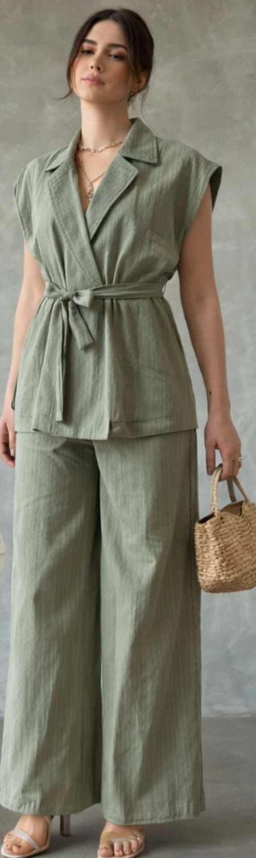 Woman wearing a green outfit with a belt, holding a straw bag against a neutral background