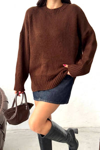 A person wearing an oversized brown ribbed knit sweater, a denim mini skirt, and black knee-high boots, holding a dark brown handbag against a plain light background.