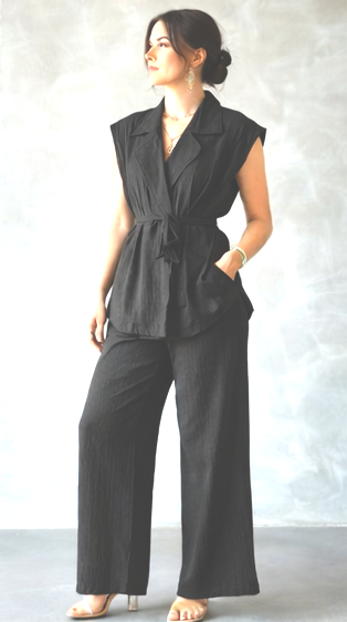 A woman models a black sleeveless wrap-style collared top paired with matching black wide-leg pants, standing against a light gray textured studio backdrop.