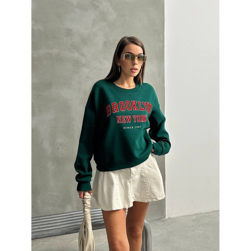 Woman wearing a green 'Brooklyn New York' sweatshirt and white skirt against a gray wall.