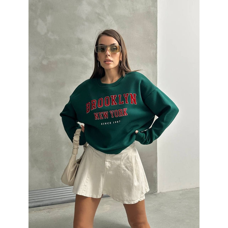 Woman wearing a green 'Brooklyn New York' sweatshirt with sunglasses and a beige bag.