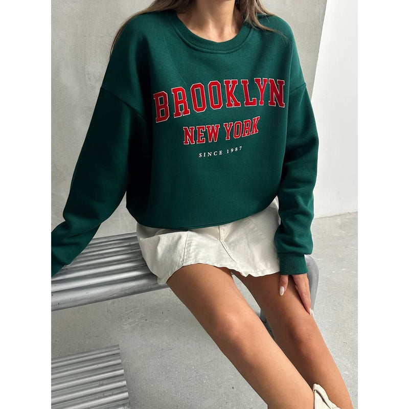 Person wearing a green sweatshirt with 'Brooklyn New York' text, sitting on a concrete surface.