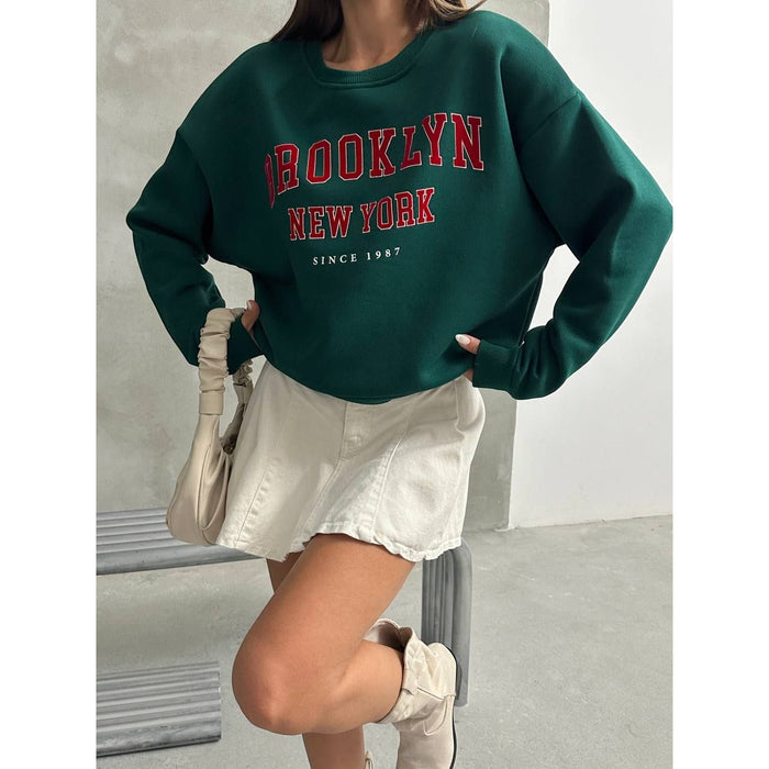 Person wearing a green 'Brooklyn New York' sweatshirt with white shorts and beige bag.