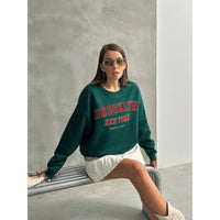 Woman wearing a green 'Brooklyn New York' sweatshirt sitting on a bench.