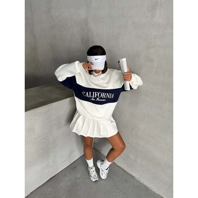 Person wearing a white and navy sweatshirt with 'California' text, white skirt, and white shoes against a gray wall.