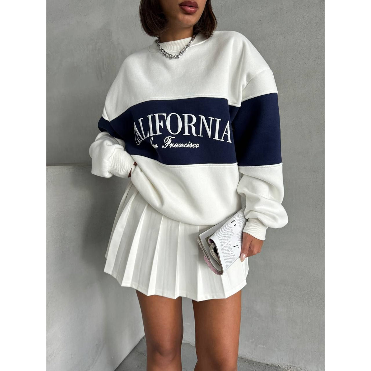 Person wearing a white and black 'California' sweatshirt with a pleated skirt against a gray background