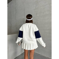 Person wearing a white and black sweatshirt with a pleated skirt against a gray wall.