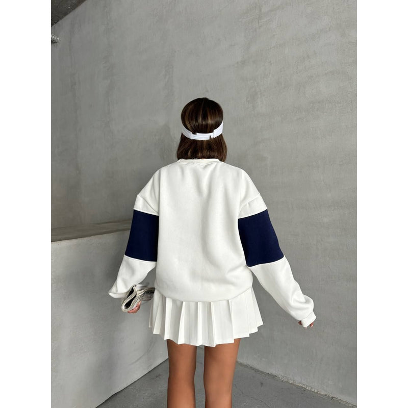 Person wearing a white and black sweatshirt with a pleated skirt against a gray wall.