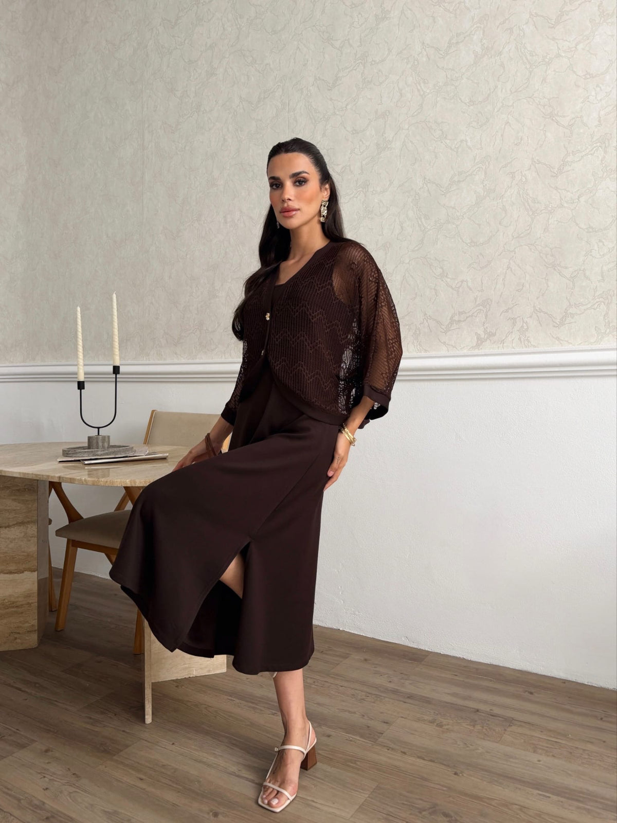 A woman models a dark brown midi dress with a sheer mesh long-sleeve top overlay in a modern indoor setting with a light patterned wall and wood flooring.
