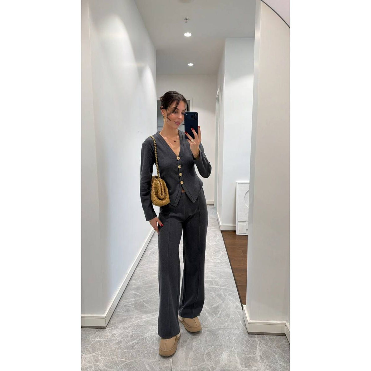 Woman taking a mirror selfie wearing a dark gray suit with a gold handbag in a hallway.