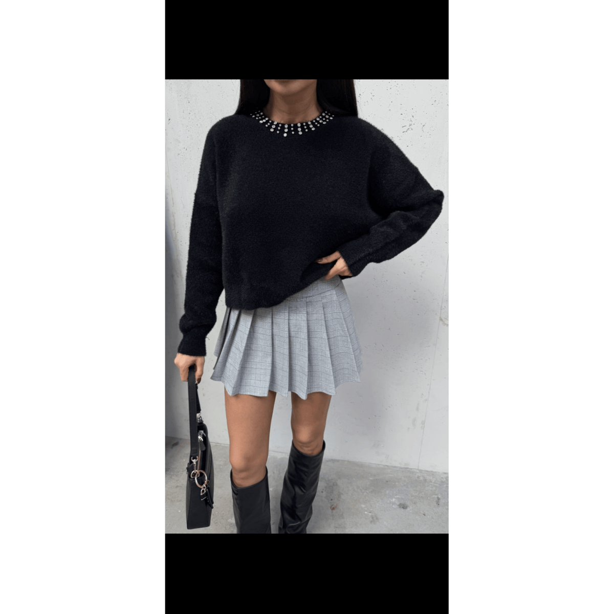 Person wearing a black sweater, gray plaid skirt, and black knee-high boots against a white wall.
