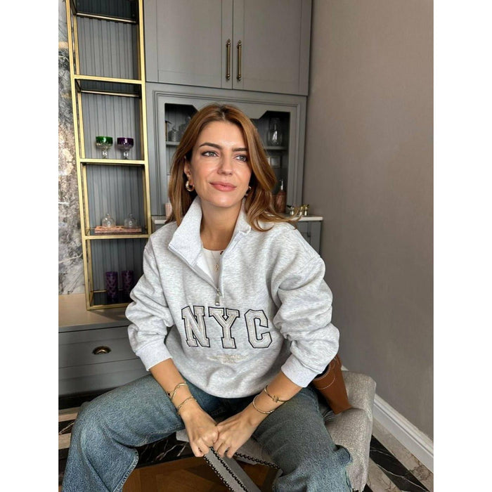 Woman wearing a white 'NYC' sweatshirt sitting on a chair indoors.