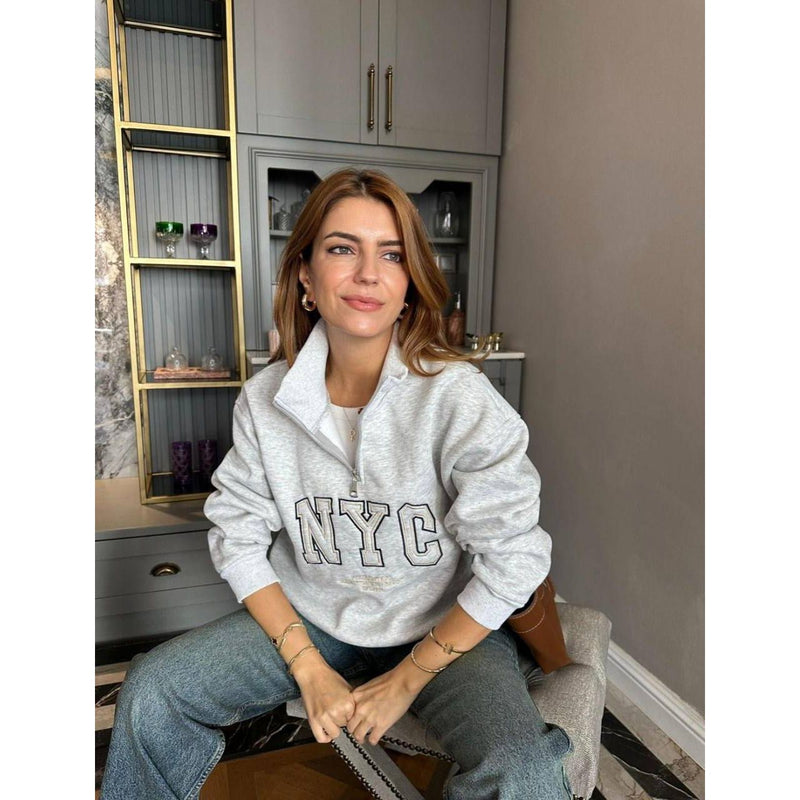 Woman wearing a white 'NYC' sweatshirt sitting on a chair indoors.