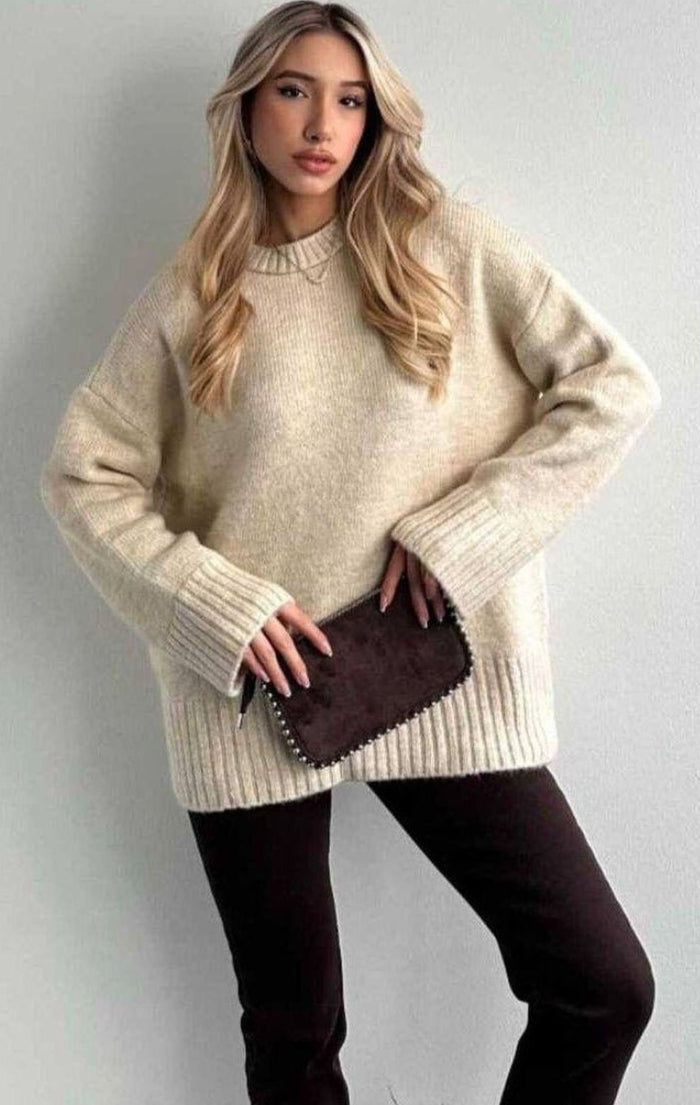 Woman wearing a beige sweater and dark pants against a plain background