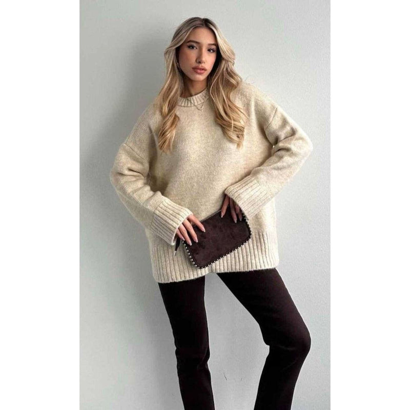 Woman wearing a beige sweater and dark pants against a plain background