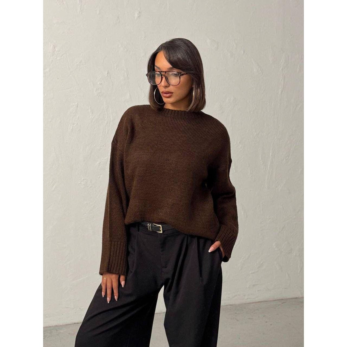 Woman wearing a brown sweater and black pants against a plain background