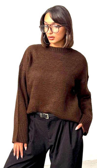 A woman wearing a dark brown knit sweater and black wide-leg trousers, standing against a plain white wall.