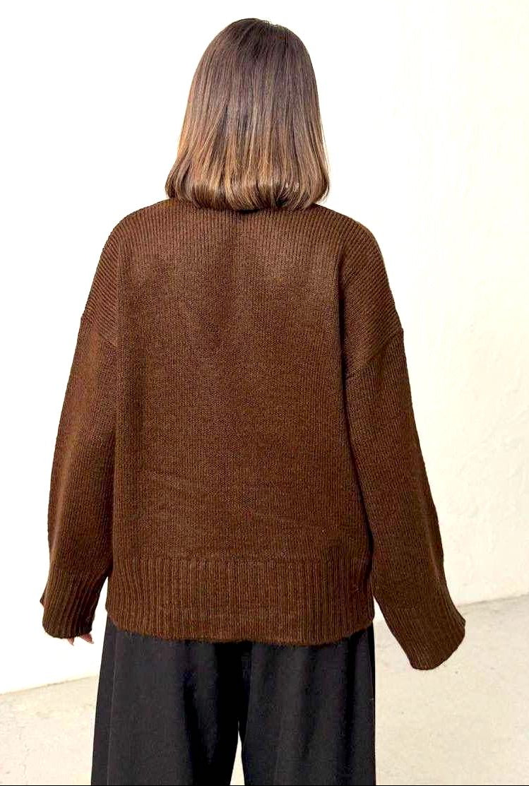 A person viewed from behind wearing an oversized brown knit sweater and dark pants against a plain off-white wall.