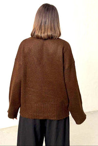 A person viewed from behind wearing an oversized brown knit sweater and dark pants against a plain off-white wall.