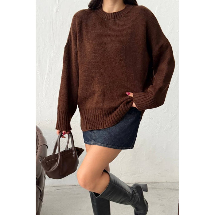 Person wearing a brown sweater, denim skirt, and black boots holding a brown handbag against a white background.