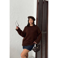 Woman wearing a brown sweater and beret holding sunglasses and a handbag against a white wall.