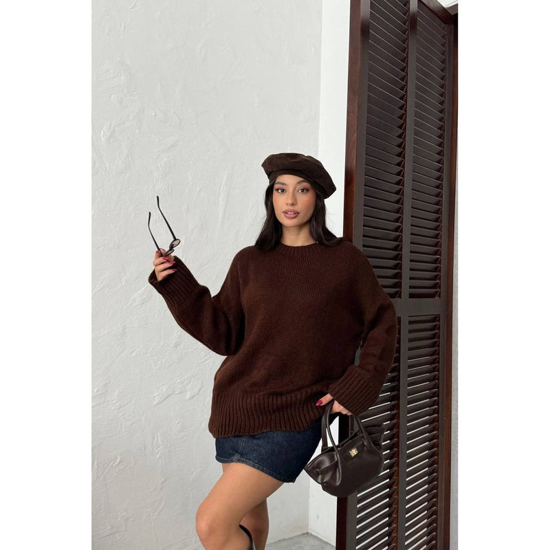 Woman wearing a brown sweater and beret holding sunglasses and a handbag against a white wall.