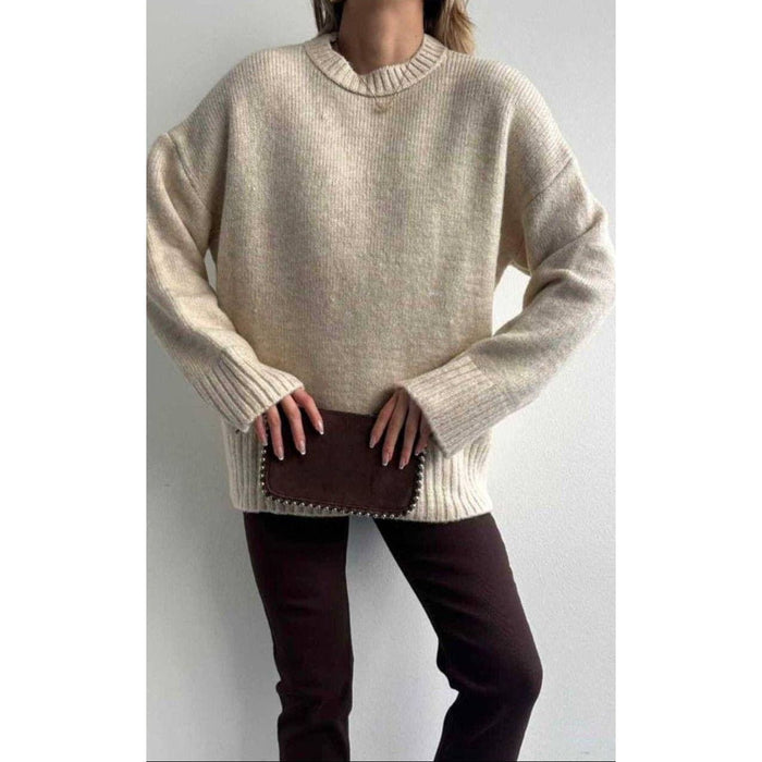 Person wearing a beige sweater and dark pants against a plain background